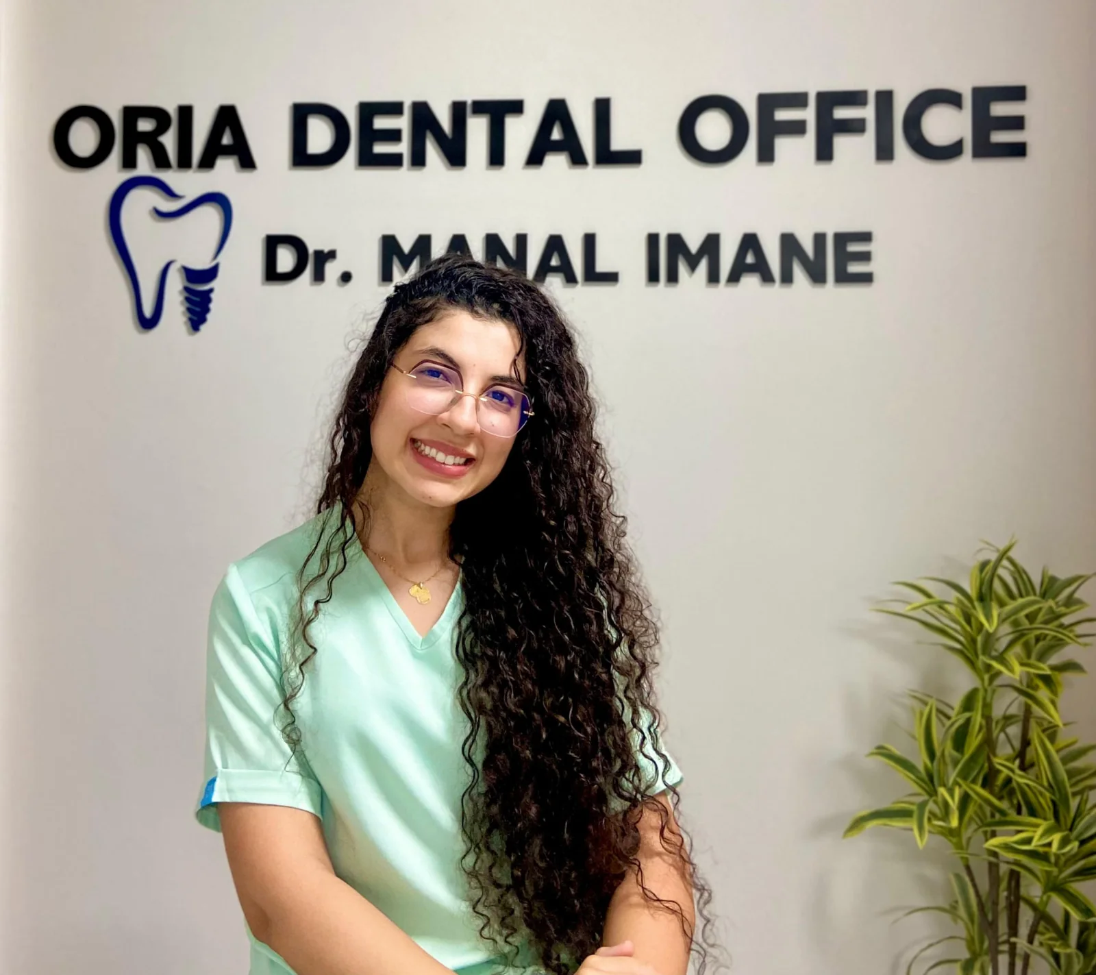 Dr Imane MANAL, chirurgien-dentiste, souriante devant le mur d'accueil du cabinet ORIA